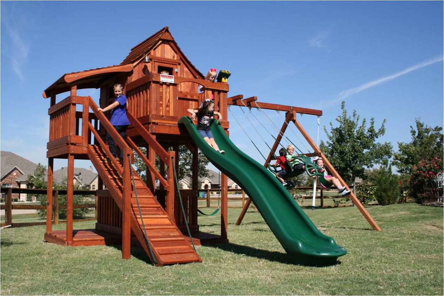 Children enjoying a large custom playset with multiple swings, slides, and climbing activities designed for growing families. Mustang swing set adventure ramp